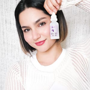 May include: A white bottle with a dropper, held up in front of a person. The bottle has a purple label with text and a graphic of a plant. The person is wearing a white sweater.