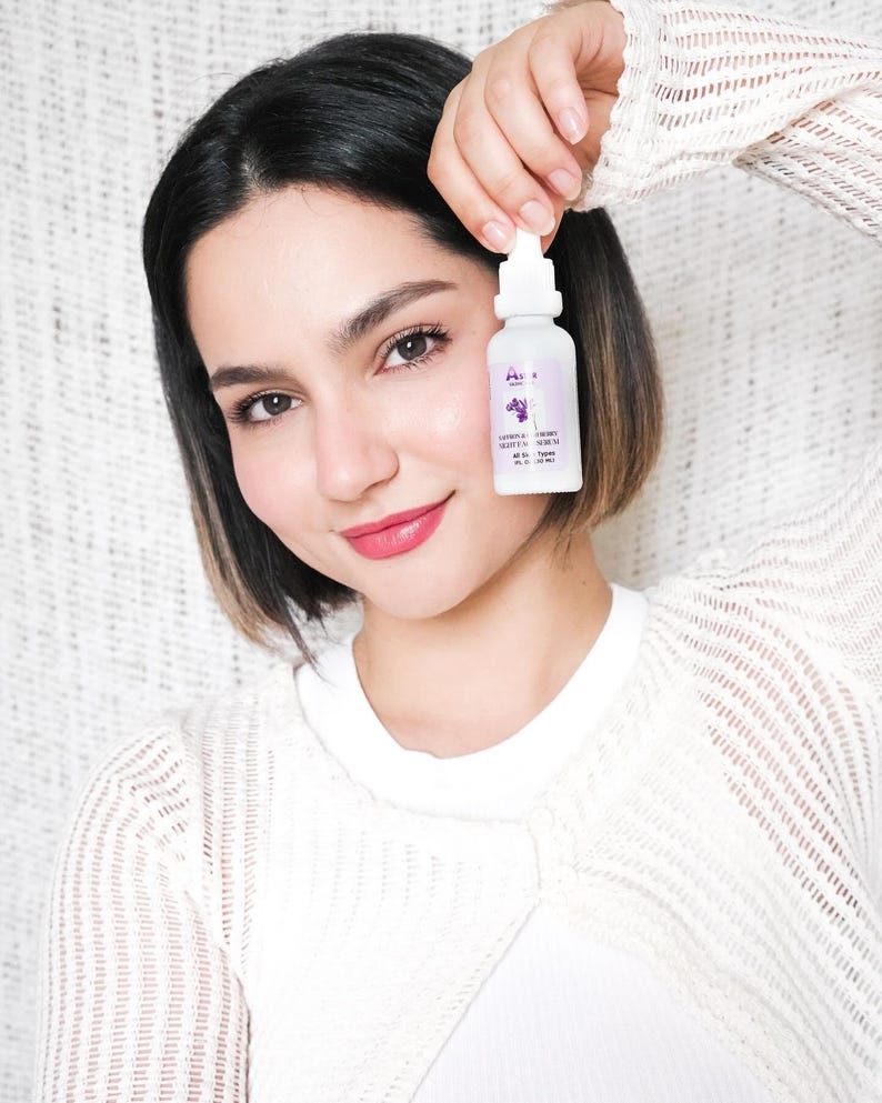 May include: A white bottle with a dropper, held up in front of a person. The bottle has a purple label with text and a graphic of a plant. The person is wearing a white sweater.