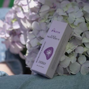 May include: A white and lavender box of Aster Face Night Serum with the words "Restorative & Radiance" and "Saffron & Gooseberry" printed on the side. The box is next to a bouquet of lavender and white hydrangea flowers.
