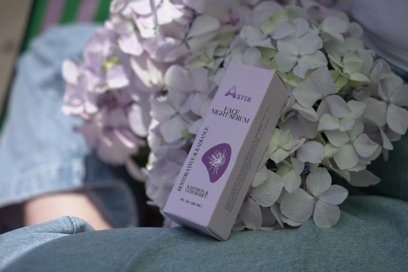May include: A white and lavender box of Aster Face Night Serum with the words "Restorative & Radiance" and "Saffron & Gooseberry" printed on the side. The box is next to a bouquet of lavender and white hydrangea flowers.