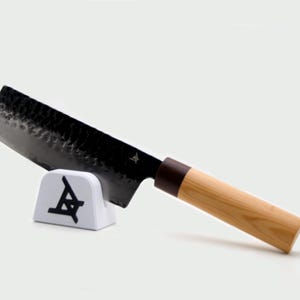 May include: A Japanese-style kitchen knife with a dark, hammered metal blade featuring a small logo near the bolster. The handle is made of light-colored wood with a dark brown ferrule. The knife rests on a small white stand with a black logo.