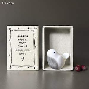May include: A small off-white matchbox measuring 4.5 x 3 cm, featuring a black ink frame and the text "Robins appear when loved ones are near" with a heart icon. Inside sits a smooth white and gray ceramic bird figurine next to two red berries.