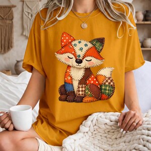 May include: A mustard yellow t-shirt featuring a central graphic of a fox designed with a patchwork quilt aesthetic. The fox illustration displays various fabric patterns including floral, plaid, and polka dots in shades of orange, brown, and blue.