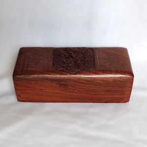 May include: A rectangular wooden box with a rich brown finish and a visible wood grain pattern. The lid features a centred, intricately carved square design with detailed floral or leaf motifs, providing a textured contrast to the smooth sides.