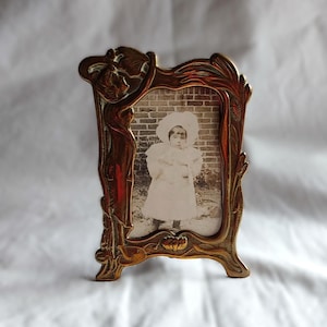 May include: A vertical brass picture frame with an ornate, flowing border design featuring raised relief details. The frame holds a black and white photograph of a child wearing a large hat and a long coat, standing against a brick wall background.