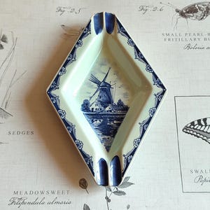 May include: A diamond-shaped ceramic ashtray featuring a cream-coloured base with a blue and white transferware illustration of a Dutch windmill by a canal. The rim is decorated with a dark blue floral pattern and includes two integrated notches.