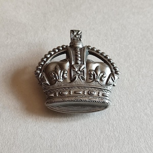 May include: A silver-toned metal crown brooch featuring intricate embossed detailing, including fleur-de-lis motifs along the base, a beaded arch design, and a central cross finial at the top. The piece has a metallic finish with dark recessed areas.