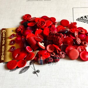 May include: A collection of assorted red buttons in various shapes, sizes, and textures, including smooth, matte, and patterned finishes. A yellow cardboard backing card with the text AERO TOP BUTTON is visible on the left side of the pile.