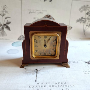 May include: A dark brown wooden mantel clock with a square gold-toned face featuring black Arabic numerals. The clock has a curved top, a gold-toned bezel, and small metal feet. The hands are positioned to indicate the time is nearly twelve o'clock.