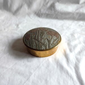 May include: An oval brass trinket box. The lid has a grey background with embossed flowers in red and brown. The box has a gold finish. The floral design is a key feature of the box.