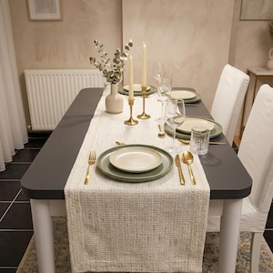 May include: A dining table set for a meal. The table has a gray top and white legs, with a cream-colored runner. The table is set with green plates, gold cutlery, and clear glassware. Two white chairs are visible.