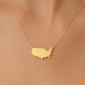May include: A gold-colored necklace featuring a pendant shaped like the United States of America. The pendant is a solid silhouette of the country's outline, suspended from a delicate chain. The necklace is worn around the neck.