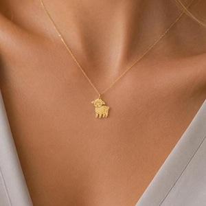 Könnte beinhalten: Eine goldfarbene Halskette mit einer zarten Kette und einem kleinen, flachen Anhänger in Form eines stilisierten Lamms. Der Anhänger hat einfache, klare Linien und eine glatte, polierte Oberfläche und hängt mittig an der dünnen Metallkette.