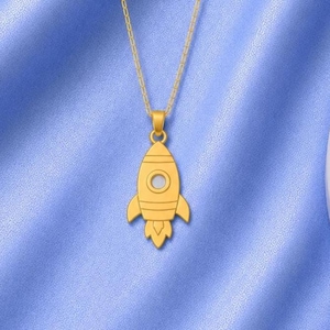 May include: A gold-toned necklace featuring a rocket ship pendant. The pendant has a smooth, polished finish with engraved horizontal lines and a circular window detail. It hangs from a delicate gold-toned chain, showcasing a simple, clean design.