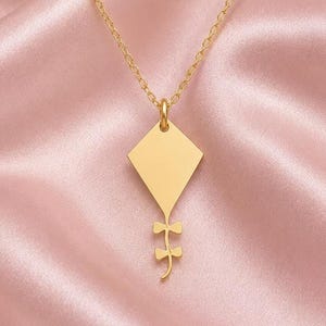 Può includere: Una collana color oro con un ciondolo a forma di aquilone dalla superficie liscia e piatta. Il ciondolo include una sezione di coda decorativa con due piccole forme simmetriche a fiocco che si estendono verso il basso dalla base.