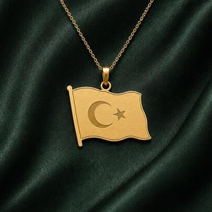 Peut inclure: Un collier pendentif doré représentant un drapeau rectangulaire aux bords ondulés. Le pendentif présente une découpe en forme de croissant de lune et d'étoile à cinq branches au centre. Il est suspendu à une fine chaîne dorée à maillons simples.