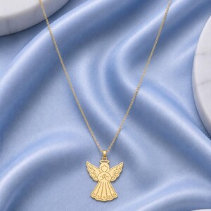 May include: A gold-toned necklace featuring a pendant shaped like a stylized angel with wings spread, a halo, and hands clasped in prayer. The angel figure has engraved line details on the wings and dress, suspended from a delicate gold-toned chain.