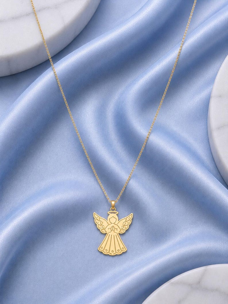 May include: A gold-toned necklace featuring a pendant shaped like a stylized angel with wings spread, a halo, and hands clasped in prayer. The angel figure has engraved line details on the wings and dress, suspended from a delicate gold-toned chain.
