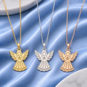 May include: Three necklaces featuring angel pendants in gold, silver, and rose gold tones. Each pendant depicts a stylized angel with wings spread, hands clasped in prayer, and a halo above the head, suspended from a matching delicate metal chain.