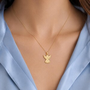 May include: A gold necklace featuring a delicate pendant shaped like an angel with outstretched wings and a halo. The pendant is suspended from a thin, polished gold chain that rests against the collarbone, showcasing a detailed, symmetrical design.