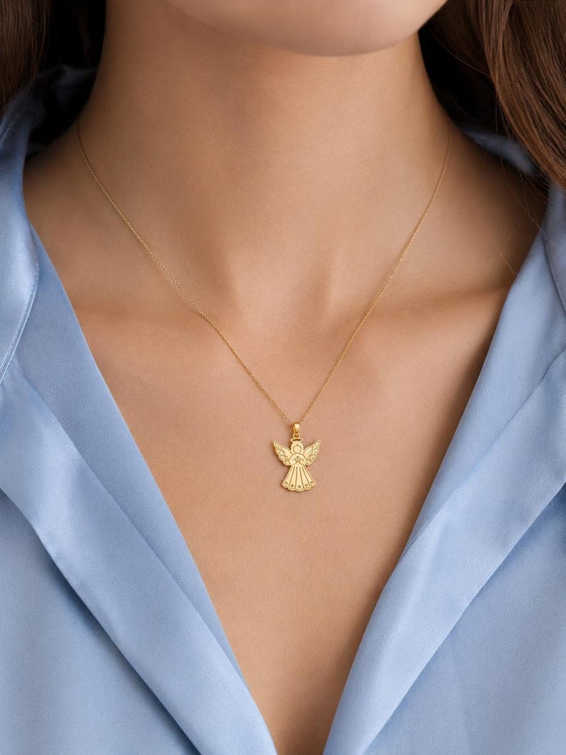 May include: A gold necklace featuring a delicate pendant shaped like an angel with outstretched wings and a halo. The pendant is suspended from a thin, polished gold chain that rests against the collarbone, showcasing a detailed, symmetrical design.