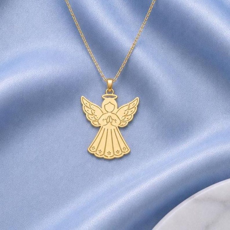 May include: A gold-toned necklace featuring a pendant shaped like an angel with outstretched wings and hands held together in a prayer position. The angel figure has etched details on the wings and a flared skirt with small star-like patterns at the hem.