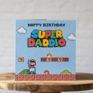 May include: A birthday card with a Super Mario Bros. theme. The card features the text "Happy Birthday Super Daddio" in a pixelated font, with a pixelated Mario character and level design on a light blue background.
