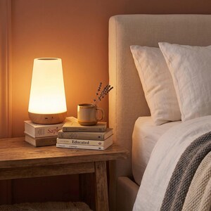 Puede incluir: Una escena de mesita de noche acogedora con una lámpara blanca y de madera, libros y una taza con lavanda. La lámpara está sobre una mesa de madera junto a una cama con ropa de cama blanca y un cabecero neutro.