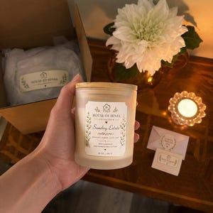 May include: A frosted glass soy candle with a wooden lid is held. The label reads "Sunday Estate" and "10 oz | 283 g". A white flower arrangement and a lit candle are in the background. A box with the brand name is also visible.