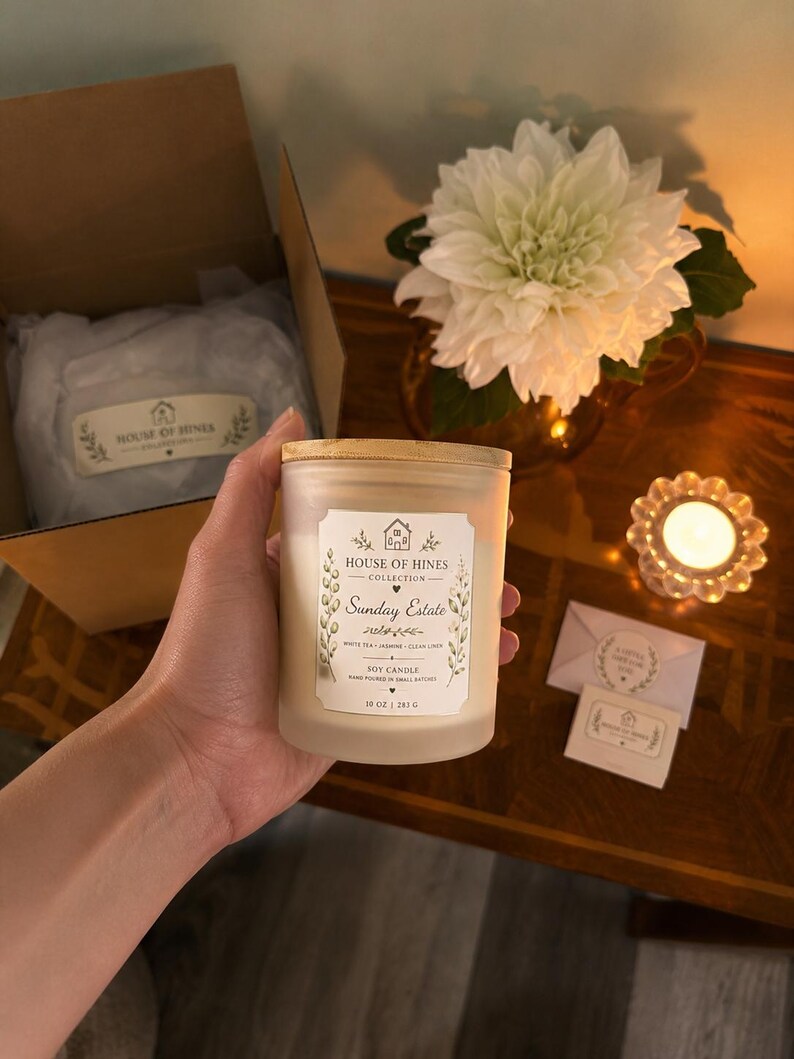 May include: A frosted glass soy candle with a wooden lid is held. The label reads "Sunday Estate" and "10 oz | 283 g". A white flower arrangement and a lit candle are in the background. A box with the brand name is also visible.