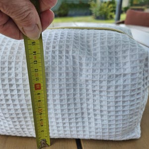 May include: A white waffle-textured fabric pouch with a gold-toned zipper closure. A yellow measuring tape is positioned vertically against the side of the pouch, indicating a height of approximately 6 inches from the base to the top edge.