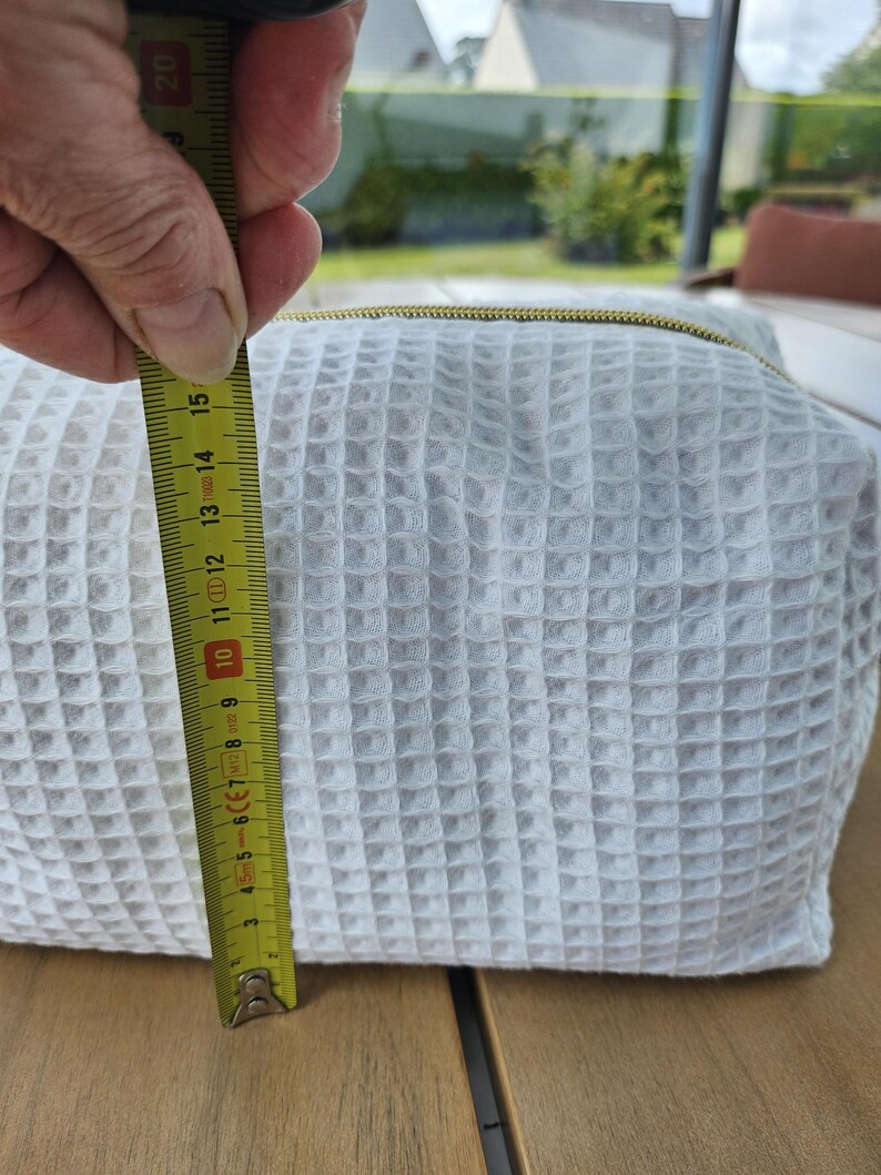May include: A white waffle-textured fabric pouch with a gold-toned zipper closure. A yellow measuring tape is positioned vertically against the side of the pouch, indicating a height of approximately 6 inches from the base to the top edge.