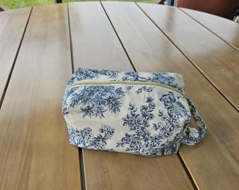 May include: A rectangular fabric pouch featuring a blue and white floral toile pattern. The bag has a gold-toned zipper closure along the top and a small fabric loop handle on the side. The exterior is made of a light-colored textile material.