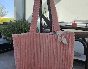 May include: A dusty rose corduroy tote bag with matching handles hangs on a dark wooden chair. The bag features a rectangular shape and a floral fabric accent. The bag is made of a soft, ribbed corduroy material.