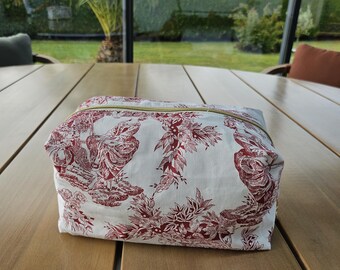 May include: A rectangular zippered pouch featuring a white fabric base with a repeating dark red toile de jouy pattern depicting pastoral scenes and floral motifs. The bag has a gold-toned metal zipper closure along the top edge.
