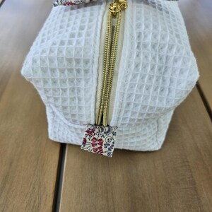 May include: A white waffle-textured fabric pouch featuring a gold-toned metal zipper closure. The zipper pull is accented with a small fabric tab in a floral pattern of red, pink, and blue on a cream background, matching the bow at the top.