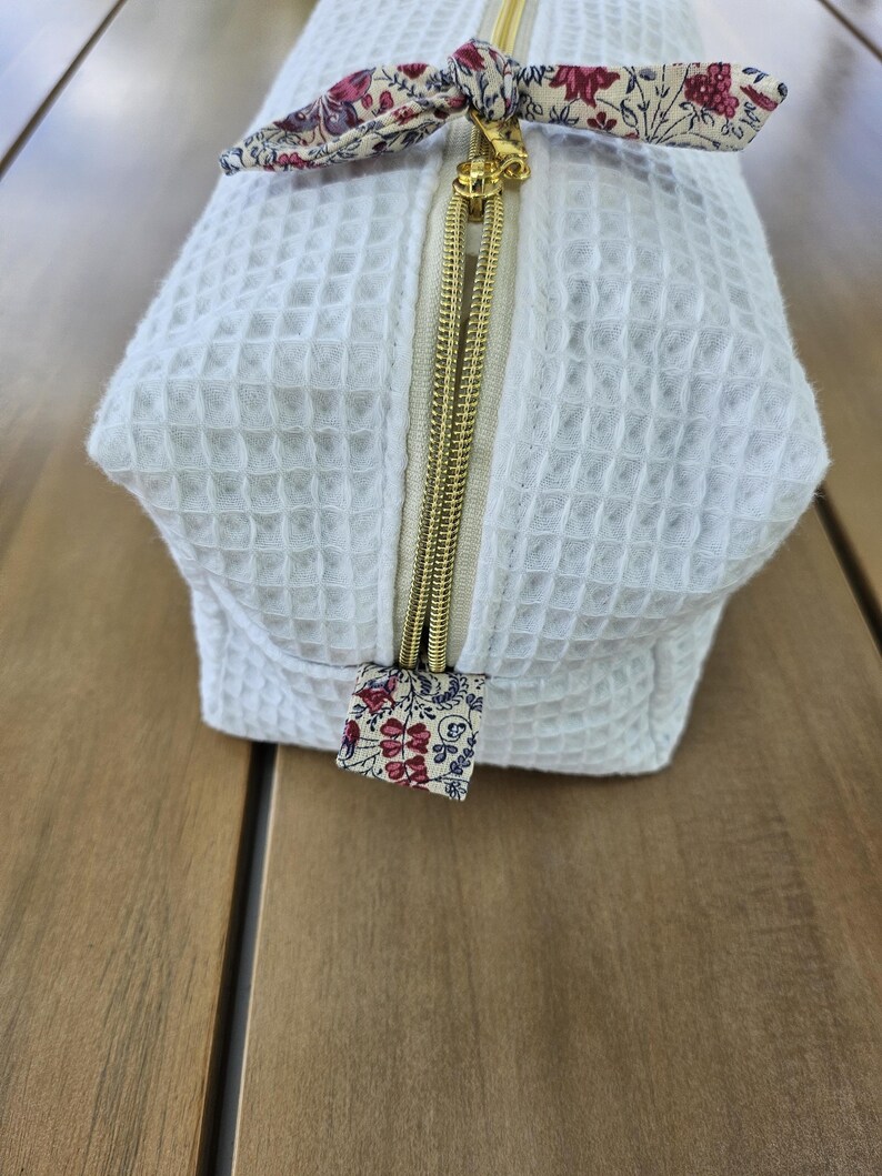 May include: A white waffle-textured fabric pouch featuring a gold-toned metal zipper closure. The zipper pull is accented with a small fabric tab in a floral pattern of red, pink, and blue on a cream background, matching the bow at the top.