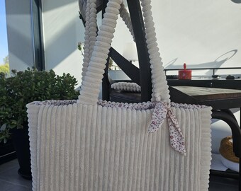 May include: A cream-colored corduroy tote bag with matching corduroy handles. The bag has a rectangular shape and a floral patterned fabric lining the top edge. A small floral ribbon is tied to one of the handles.