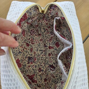 May include: A white waffle-textured fabric pouch with a yellow zipper closure. The interior is lined with a beige fabric featuring a dense floral pattern in shades of red, burgundy, and dark blue, including two internal side pockets.