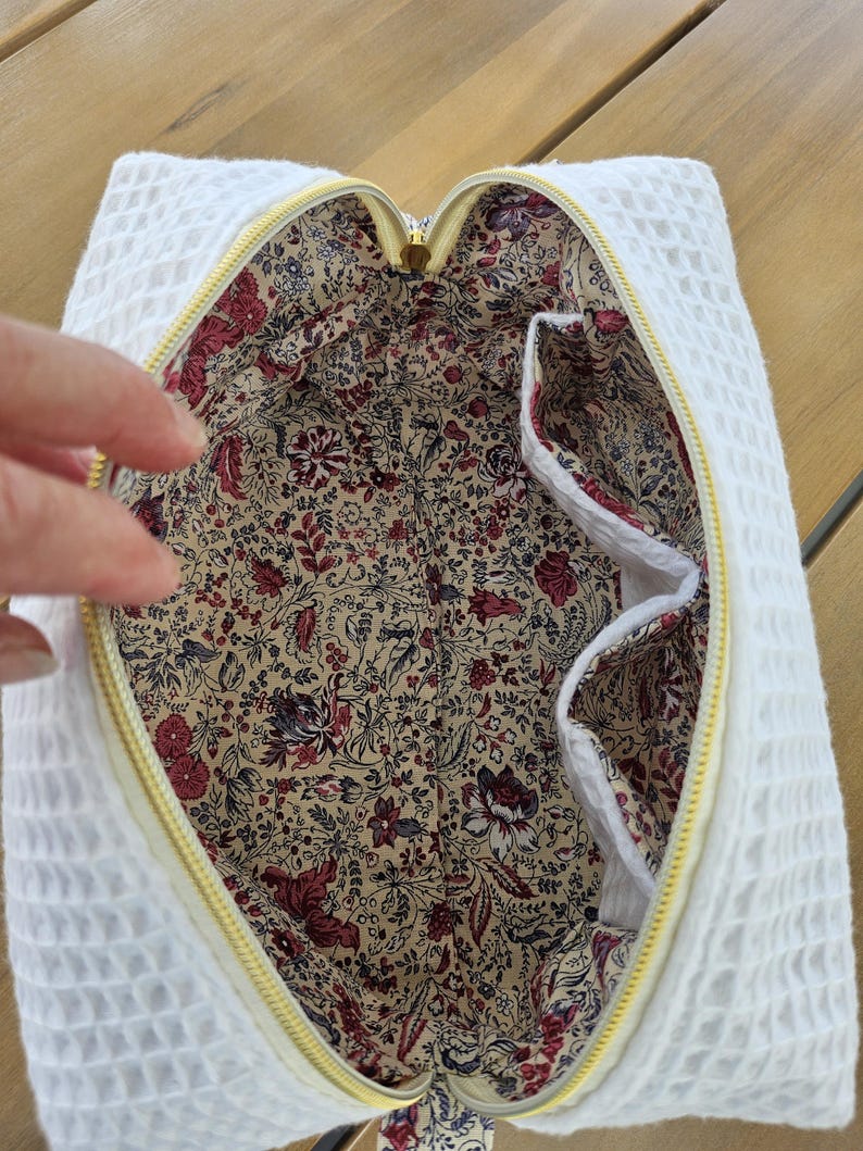 May include: A white waffle-textured fabric pouch with a yellow zipper closure. The interior is lined with a beige fabric featuring a dense floral pattern in shades of red, burgundy, and dark blue, including two internal side pockets.