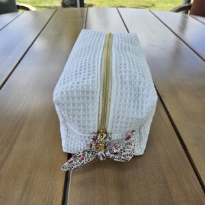 Puede incluir: Un neceser rectangular de color blanco con textura de gofre y cierre de cremallera metálica dorada. El tirador de la cremallera está adornado con un pequeño lazo de tela de color crema con un delicado estampado floral en rojo, azul y verde.