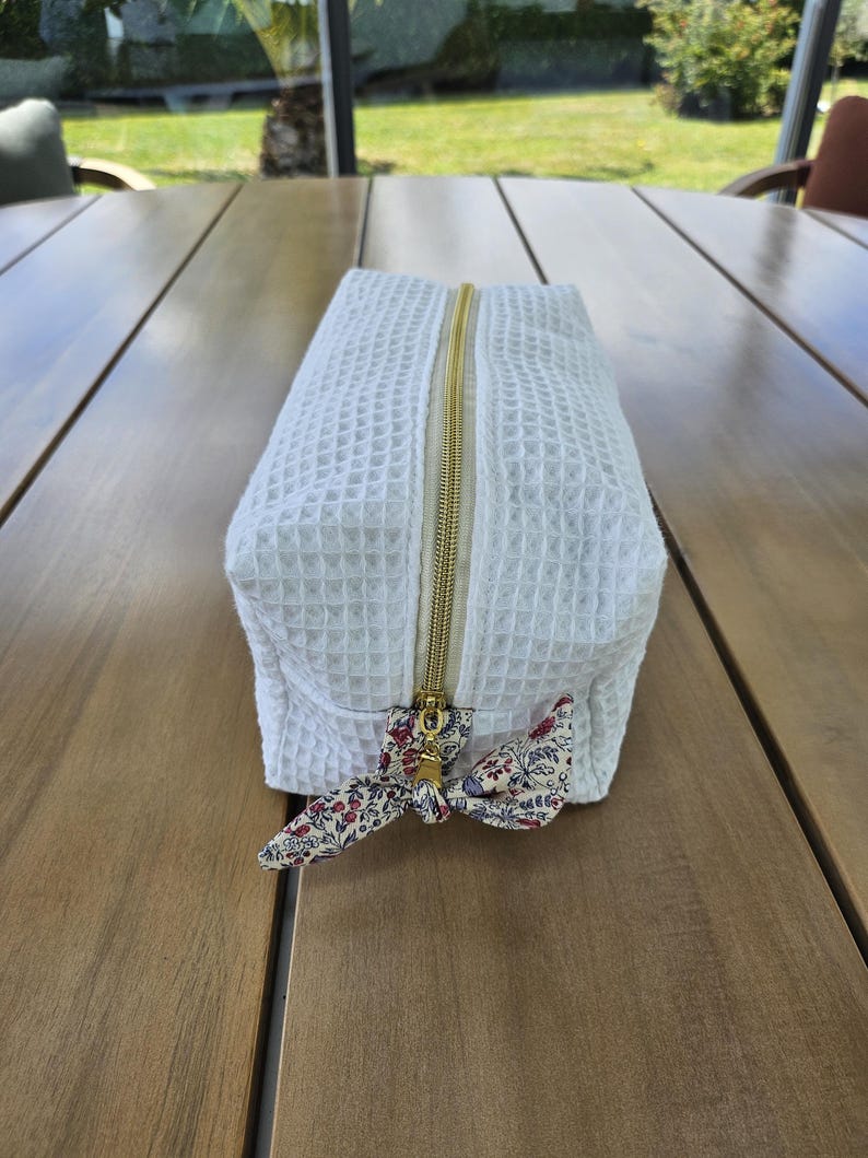 May include: A rectangular white waffle-textured toiletry bag featuring a gold-toned metal zipper closure. The zipper pull is accented with a small, tied fabric bow in a cream color with a delicate red, blue, and green floral pattern.