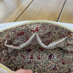 May include: A white waffle-textured zippered pouch with a gold-tone zipper. The interior is lined with a cream-colored fabric featuring a dense floral pattern in shades of red, burgundy, blue, and green, including two internal slip pockets.