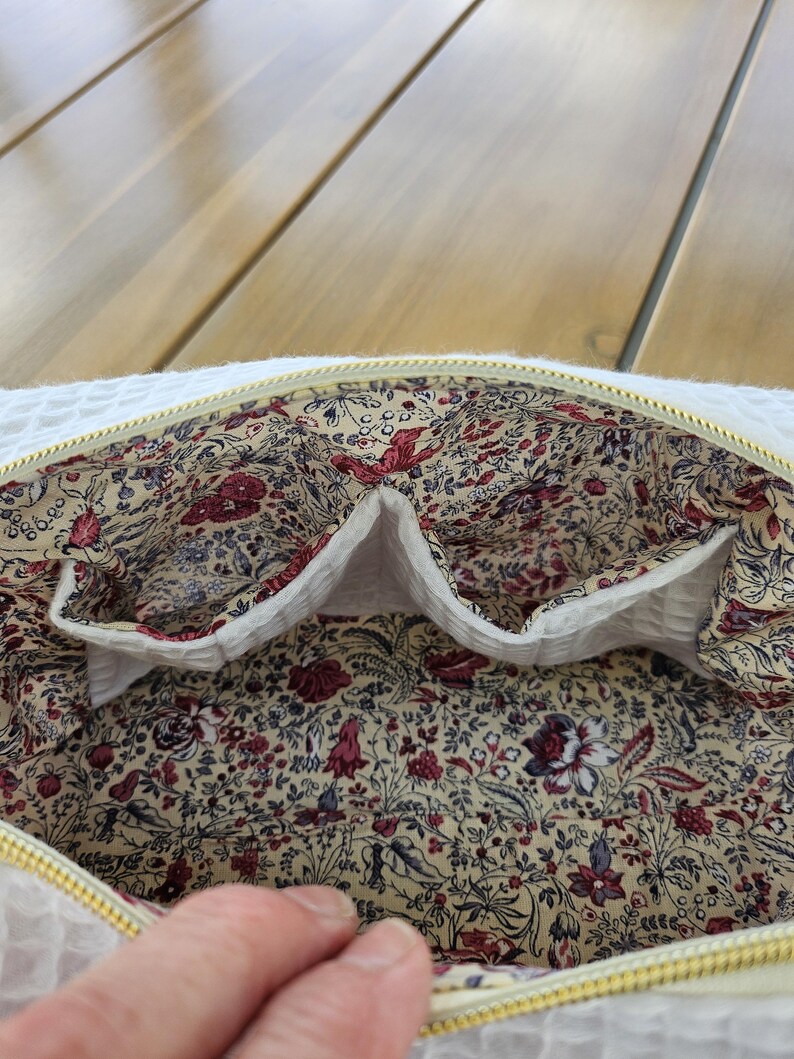 May include: A white waffle-textured zippered pouch with a gold-tone zipper. The interior is lined with a cream-colored fabric featuring a dense floral pattern in shades of red, burgundy, blue, and green, including two internal slip pockets.