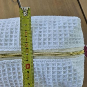 May include: A white waffle-textured fabric pouch featuring a gold-toned zipper closure. A yellow measuring tape is positioned vertically across the center of the pouch, indicating a height of approximately 6 inches for the main body of the item.