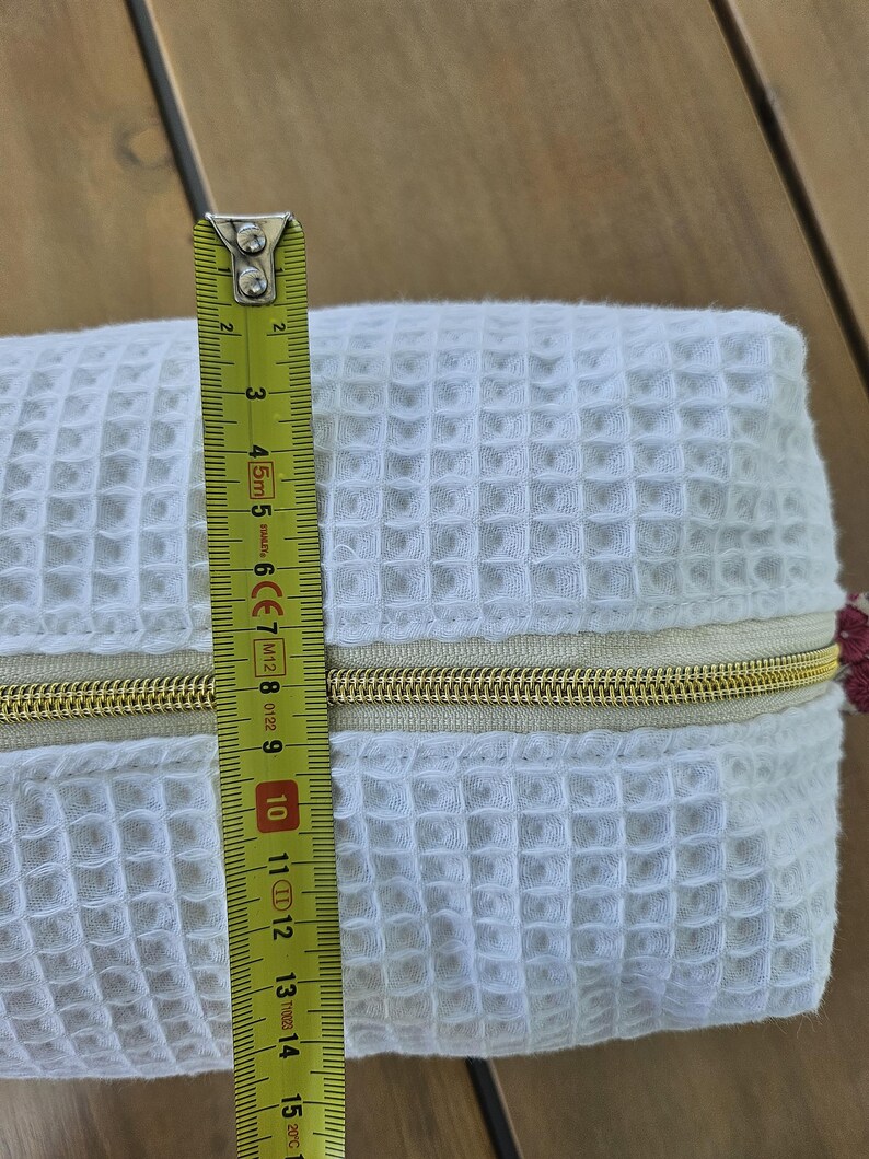 May include: A white waffle-textured fabric pouch featuring a gold-toned zipper closure. A yellow measuring tape is positioned vertically across the center of the pouch, indicating a height of approximately 6 inches for the main body of the item.
