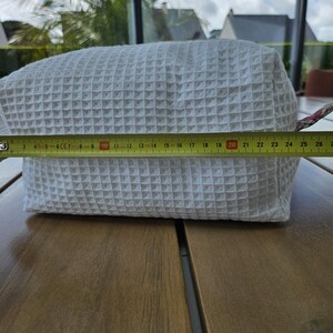 May include: A white rectangular pouch with a textured waffle-weave pattern. A yellow measuring tape is positioned across the front, indicating a length of approximately 10.25 inches from the left edge to the right side of the fabric pouch.