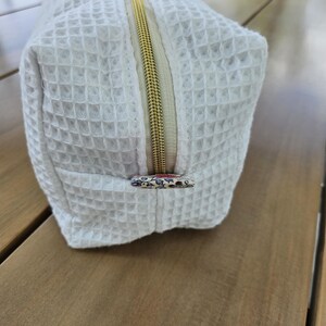 May include: A white rectangular toiletry bag with a waffle-textured fabric surface. The bag features a gold-toned metal zipper closure and a small floral-patterned fabric pull tab at the end of the zipper, matching the floral fabric knot on top.