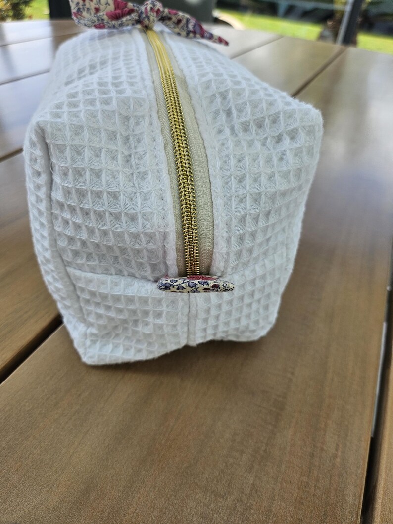 May include: A white rectangular toiletry bag with a waffle-textured fabric surface. The bag features a gold-toned metal zipper closure and a small floral-patterned fabric pull tab at the end of the zipper, matching the floral fabric knot on top.
