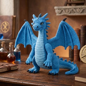 Könnte beinhalten: Ein hellblauer gehäkelter Drache mit großen Flügeln, der auf einer Holzoberfläche steht. Der Drache hat ein detailliertes, strukturiertes Design und ist von Büchern, Flaschen und einem Kompass umgeben. Die Flügel des Drachen sind ausgebreitet, und er hat einen freundlichen Ausdruck.